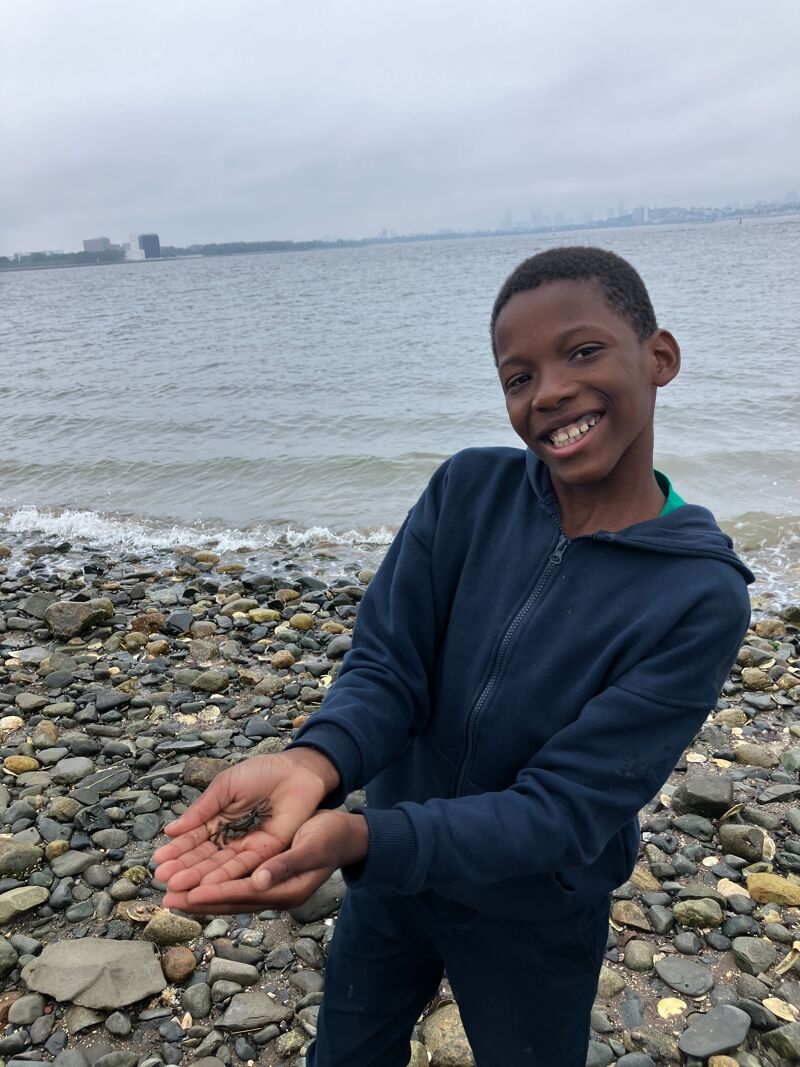 The image shows a young boy smiling at the camera while standing on a rocky beach. He is holding something small in his hands. The background features a body of water, a cloudy sky, and some buildings in the distance. The boy is wearing a blue hoodie and dark pants.